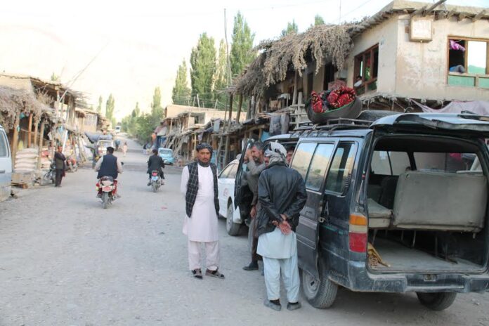 Men stand beside a dusty road with a blue van loaded with goods in a rural market.