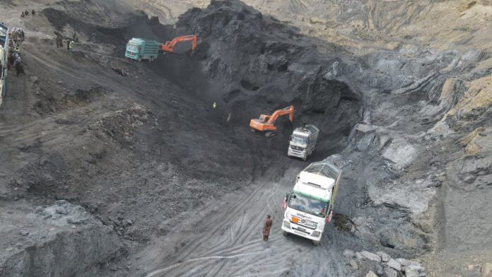 Open-pit mining scene with an orange excavator loading a truck in a deep, dark coal pit; several trucks and workers in reflective vests along the dirt road.